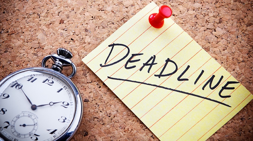 The word "deadline" written on a post-it note tacked to a cork board with a clock in the foreground.