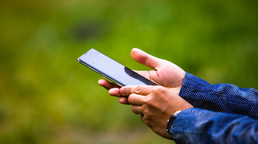 Close-up of a person holding and using a smartphone outdoors, with a blurred green background.