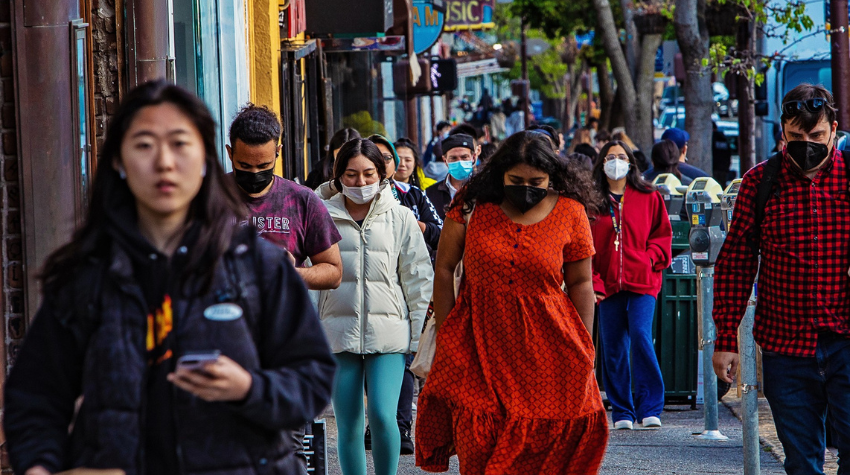 Wide shot of a crowded street in Berkeley with many people walking towards the camera, some masked some unmasked.