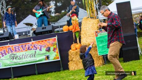 Father dances with toddler daughter outdoors at park in front of a stage