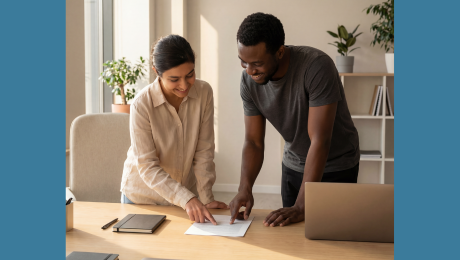 Two people smiling and standing next to each other at a desk while pointing to a piece of paper