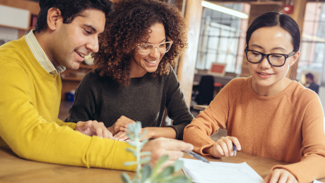Three people sit at a table smiling while one shows the other two where to sign on the paper in front of them.