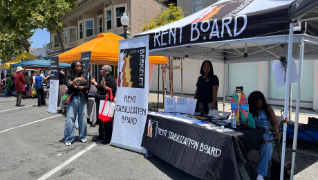 Rent Board employees sit and stand behind a table and under a Rent Board tent. One employee speaks with a community member off to the side.