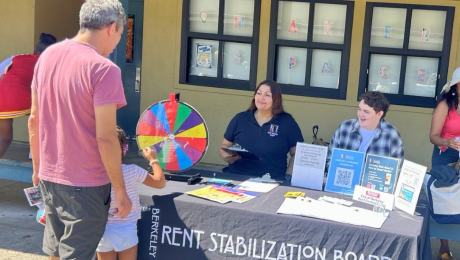 Two Berkeley Rent Board housing counselors sit at a table of resources and are visited by a father and daughter.